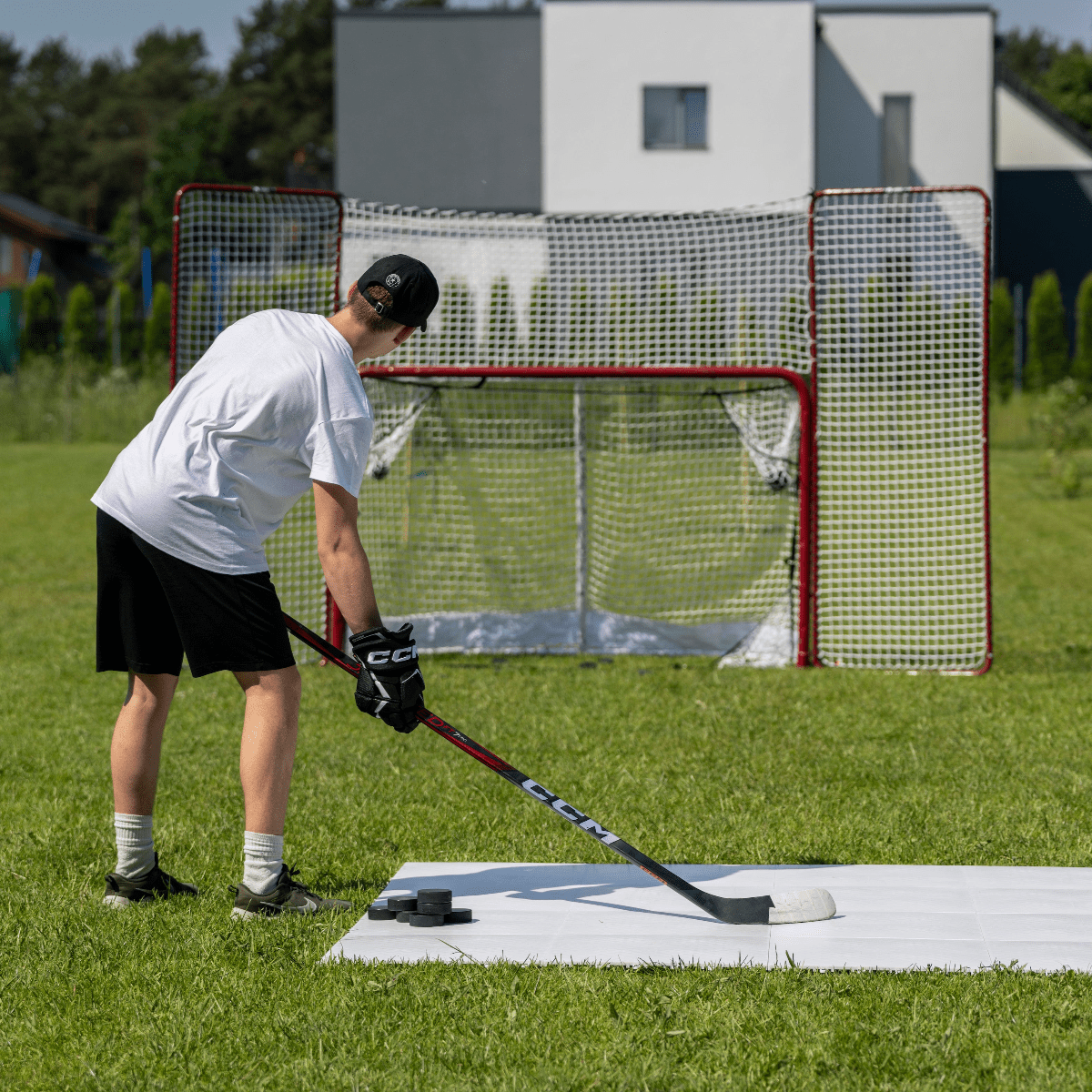 En spelare övar skott med en hockeyklubba framför ett hockeymål i gräset, med puckar framför sig. Perfekt för träning hemma.
