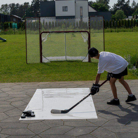 Backyard home hockey training setup with a roll-up shooting pad, pucks, and a full-size goal with backstop
