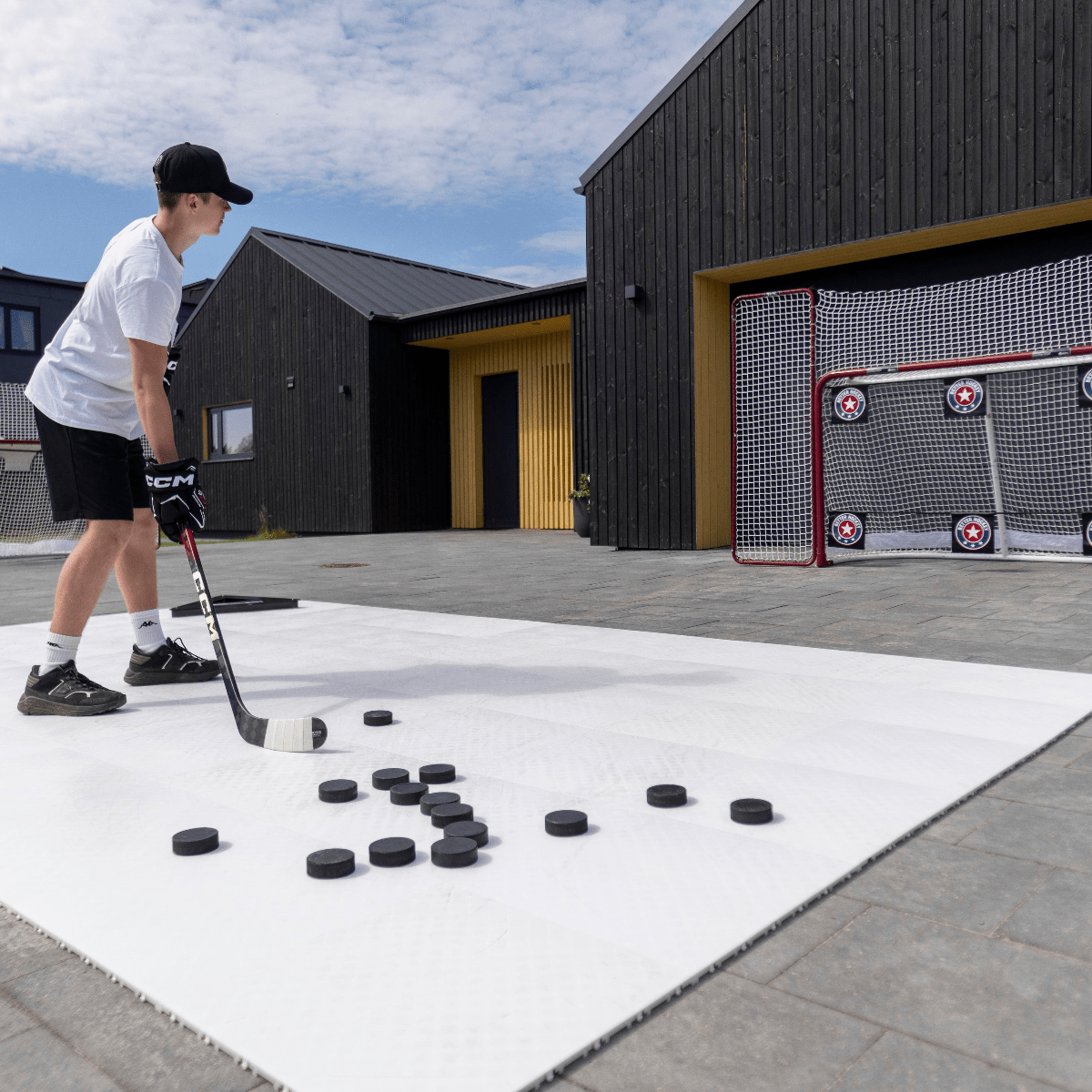 En spelare skjuter puckar mot ett hockeymål med skyddsnät, omgiven av pucks på en vit yta utomhus.