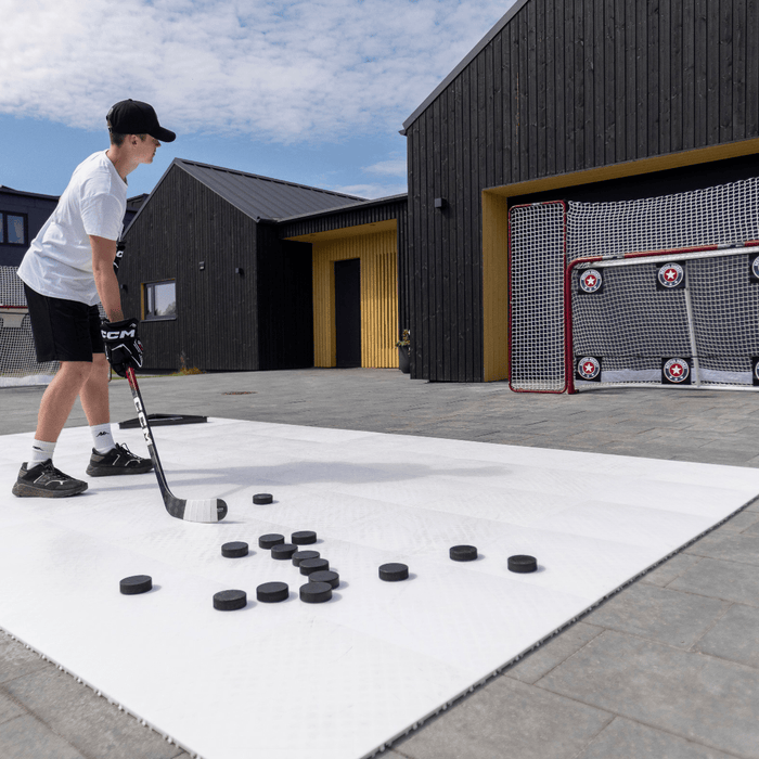 En spelare skjuter puckar mot ett hockeymål med skyddsnät, omgiven av pucks på en vit yta utomhus.