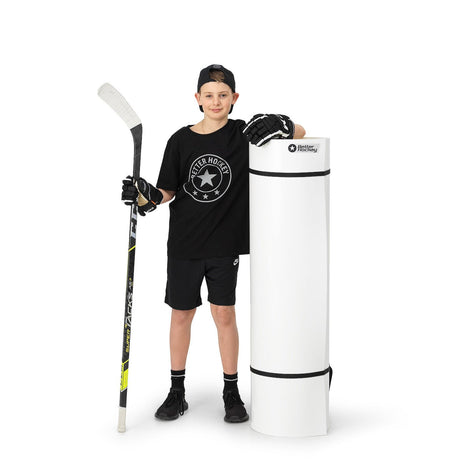 Ung hockeyspelare i svart tröja och shorts står bredvid en rullad skottplatta, redo att träna sin hockeypassning.