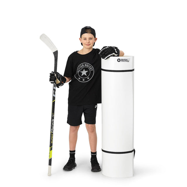 Ung hockeyspelare i svart tröja och shorts står bredvid en rullad skottplatta, redo att träna sin hockeypassning.
